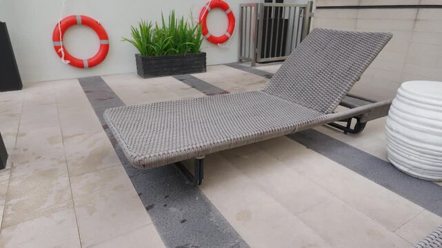 Comfortable wicker sun lounger by the outdoor swimming pool deck with lifebuoys.