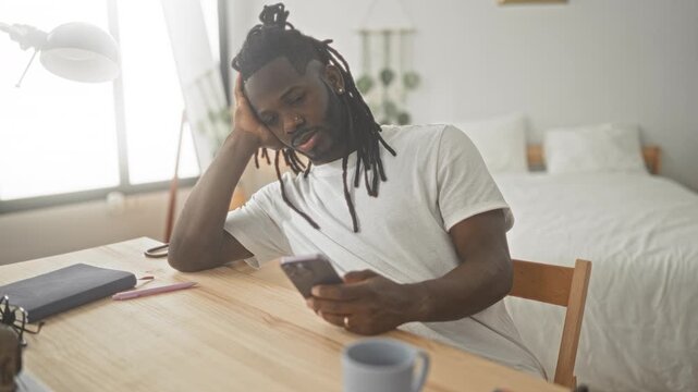 Man holds smartphone with one hand, head resting on palm, slouched at wooden table beside bed in studio; tired distraction.