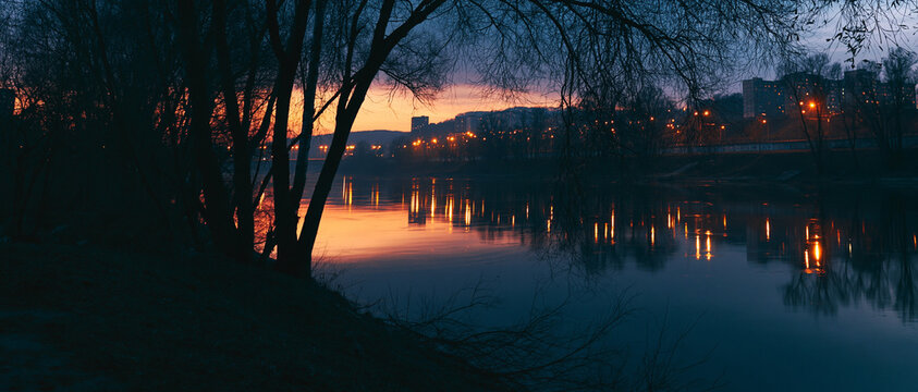 Tranquil river at dusk reflecting city lights and silhouettes of trees  