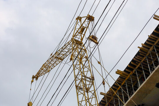 A yellow tower crane rises beside the reinforced concrete building frame under an overcast sky. Steel cables and crane jib are actively involved in high-altitude work on the construction site.