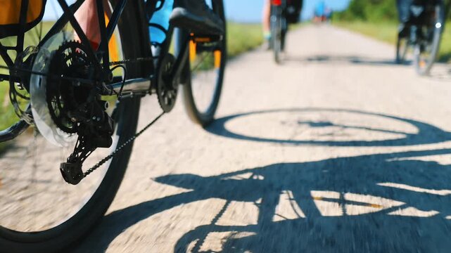 Low-angle close-up of a cyclist pedaling legs and bike drivetrain on a gravel section of the EuroVelo 10 route in Denmark, with other riders ahead and a bike shadow on a sunny summer day