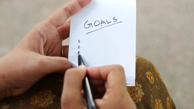 Woman writing numbered list under goals header on white notepad while sitting outdoors in daylight