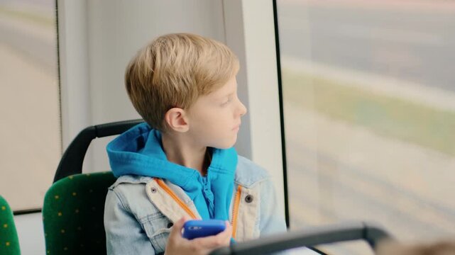 Three-quarter profile of young blond boy seated on public bus holding blue smartphone at chest level while turning head to gaze out window, expression shifts from neutral to slight smile indicating
