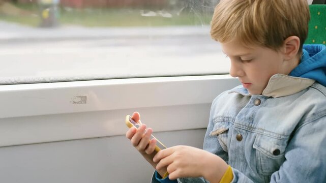Side-profile close-up of young school-age boy seated by window on public transport, holding yellow-cased smartphone with both hands and tapping or scrolling with focused downward gaze