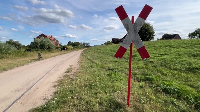 Unpaved rural road through green countryside with a red and white railway crossing warning sign