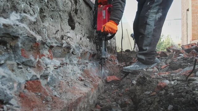 A worker uses a red electric jackhammer to remove an old concrete or stone wall base. Properly prepare the foundation for repairs.