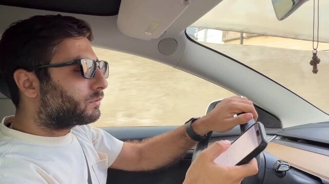Side view of a distracted bearded young man using a smartphone while driving a modern car on a city road. Dangerous behavior behind the wheel. Stock video with model release