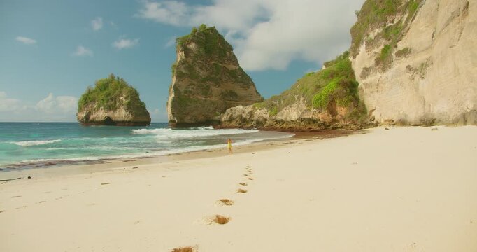 Woman in dress dances on white sand beach near large cliffs and turquoise ocean