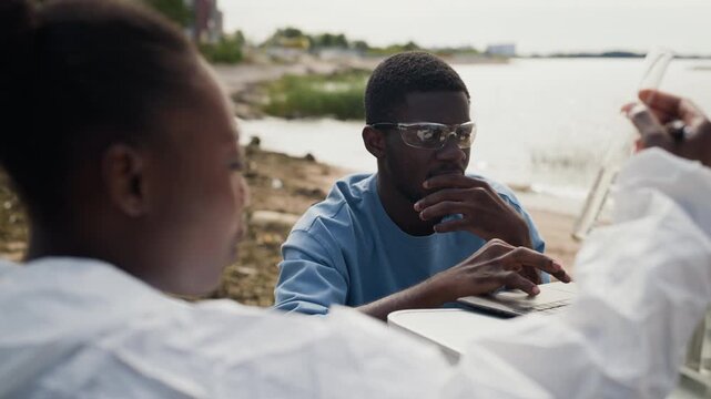 African American young adult scientists analyzing water samples and logging data into laptop outdoors. Over-shoulder view ideal for ecology reports, water contamination campaigns, or research