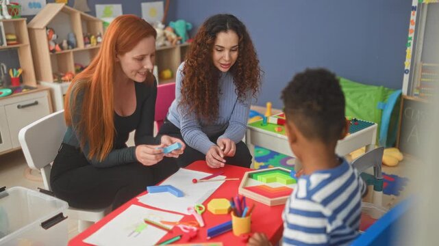 Two women teachers guide a boy placing colorful wooden blocks and stacking hexagon pieces on a play table in a school building; nurturing learning.