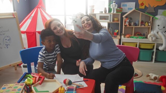 Woman taking selfie with smartphone while boy draws with crayon at a table in preschool building; playful learning.