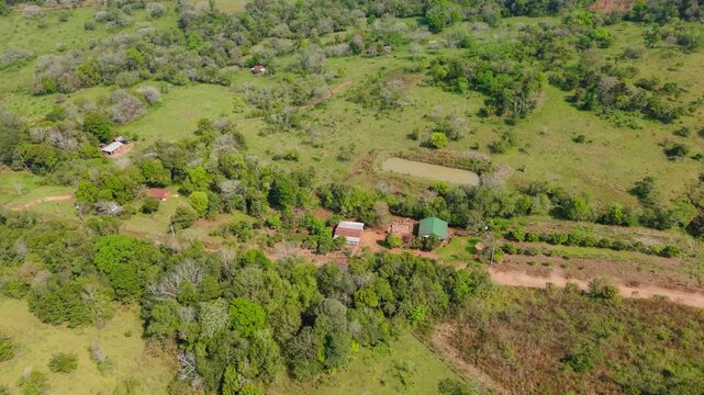Aerial view captures a car racing on the winding dirt road amid dense greenery and scattered rural buildings in Nueva Alborada. Sunlit countryside forms a vivid backdrop for the high-speed WRC rally.