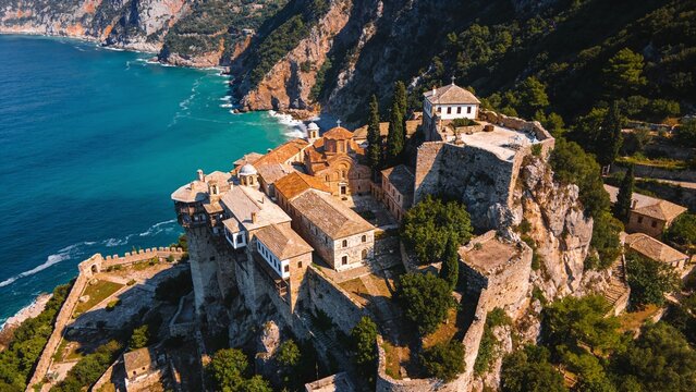 High-angle drone photograph of the Holy Skete of Saint Anna in the Monastic State of Mount Athos, nestled among rugged mountain cliffs above the Aegean coastline.