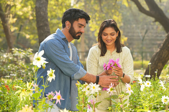 Beautiful indian young couple admiring and holding pink flowers together in a lush green park, enjoying a romantic outdoor moment surrounded by nature.