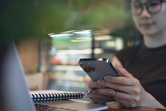 Young asian woman using smartphone while remote working on laptop with notebook on desk in cafe, modern workspace, online business lifestyle, freelance at work concept