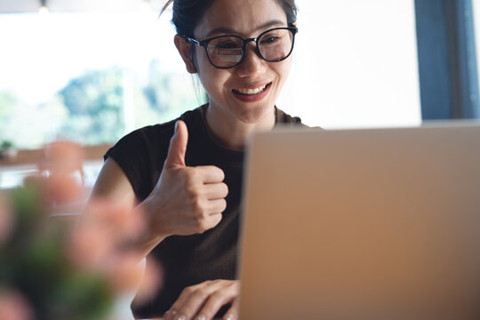 Smiling woman with glasses showing thumbs up while using laptop for video call, online meeting in modern workspace, positive remote work and success concept.