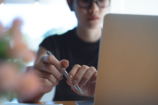 Asian woman holding pen, thinking while working on laptop at home office, calm and focused in soft natural light, modern remote work concept.