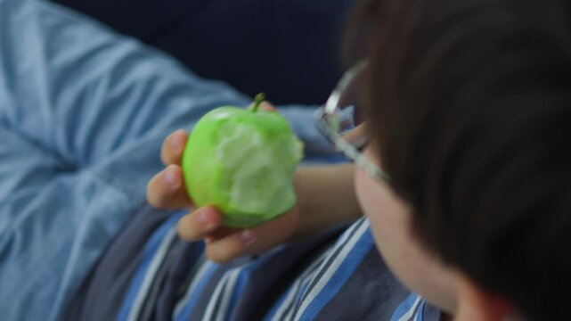 cute young boy with glasses eating a fresh green apple. Healthy childhood nutrition, daily vitamin snack, and casual indoor lifestyle portrait of a kid enjoying organic fruit.