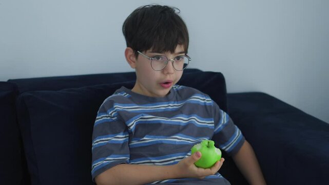 cute young boy with glasses eating a fresh green apple. Healthy childhood nutrition, daily vitamin snack, and casual indoor lifestyle portrait of a kid enjoying organic fruit.