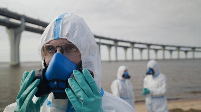 Young adult Caucasian ecologist wearing hazmat suit putting on respirator mask outdoors. Ideal for ecological disaster documentaries, biohazard research, and safety campaigns
