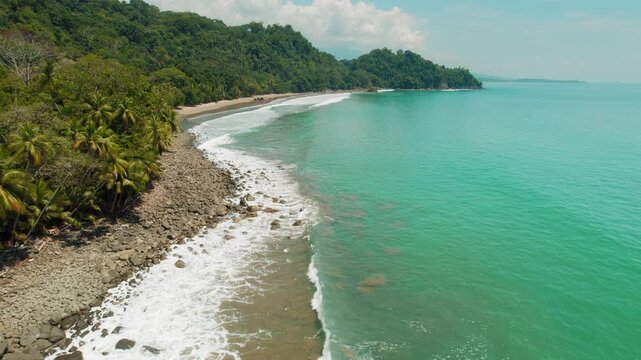 Drone flying over remote rocky bay with jungle hills and calm turquoise ocean waves