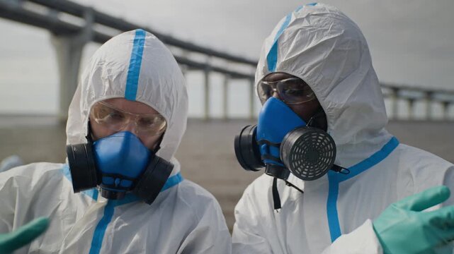 Multiethnic ecologists wearing hazmat suits analyzing disaster data on laptop by polluted coast. Footage serves environmental science documentaries and pollution crisis campaigns