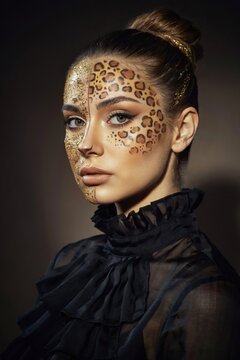 A woman with elegant makeup featuring leopard spots and glitter on her face, wearing a high-neck black ruffled blouse and her hair styled in a bun.