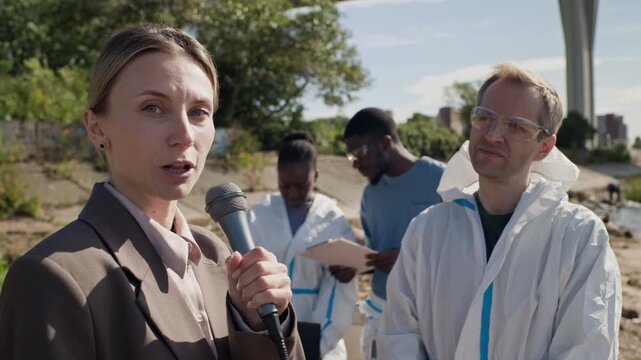 Portrait of young adult Caucasian female reporter speaking to camera and interviewing scientist in hazmat suit outdoors. Clip suitable for environmental journalism and pollution news reports