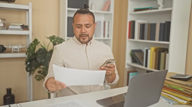 Man holding smartphone and document while reading papers at a home office desk; focused productivity.