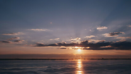 Fototapeta premium Sunset over frozen lake surface with clouds and sun rays beaming.