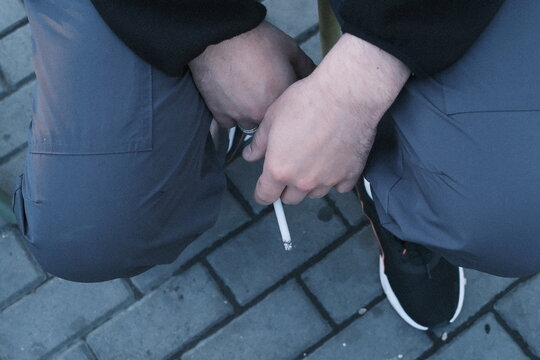 Man in casual grey pants sits on a paved outdoor surface while holding a single lit cigarette between his fingers during the quiet evening time of day