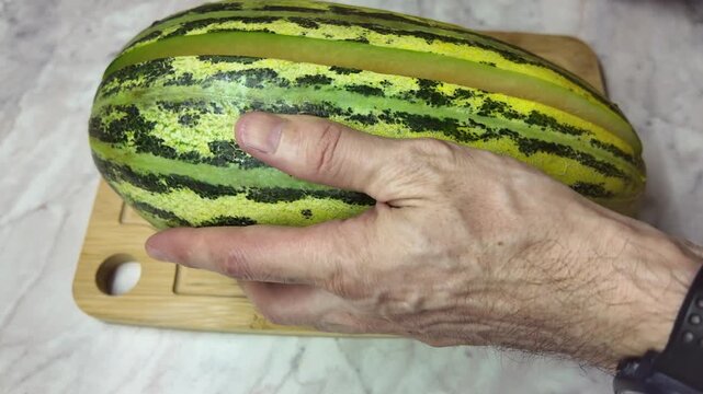 Male hands holding a large knife and cutting a fresh, ripe melon in half lengthwise on a wooden cutting board in the kitchen.