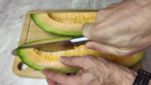 Male hands using a sharp knife to cut a fresh and juicy yellow melon into slices on a bamboo cutting board. Healthy food preparation concept