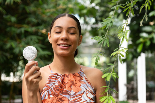 Young woman smiling with closed eyes, enjoying a cool breeze from a personal portable fan outdoors