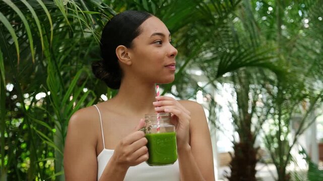 Happy young woman enjoying a healthy green detox smoothie surrounded by lush tropical plants