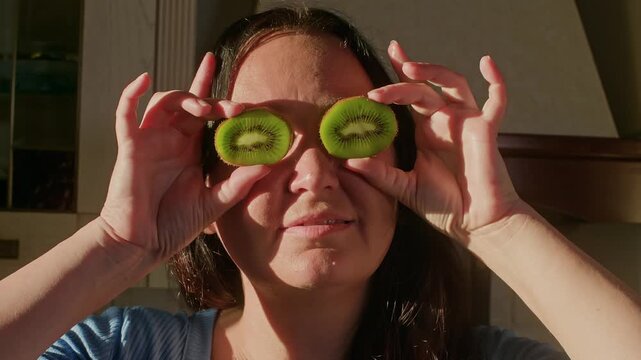 Woman holds two kiwi fruit slices over eyes like eyeglasses, playful pose captured with natural window lighting, minimal camera movement in bright interior setting.