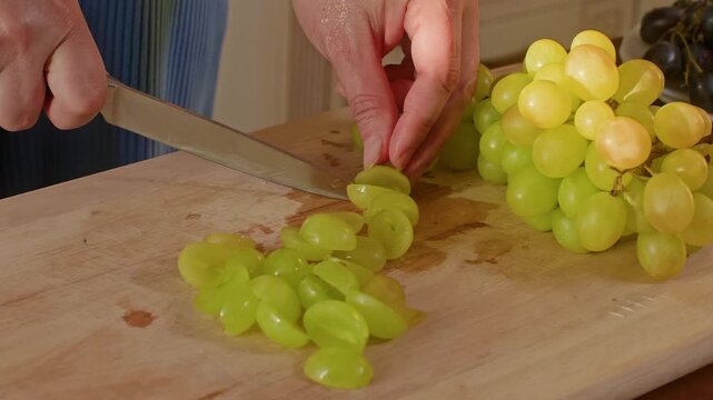Person cuts fresh green grapes with knife on wooden board in bright kitchen setting, demonstrating food preparation technique with close up camera work.