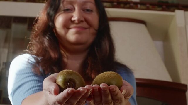 Woman in blue shirt presents two fresh kiwi fruits in her hands while smiling at camera in domestic interior setting with natural lighting.