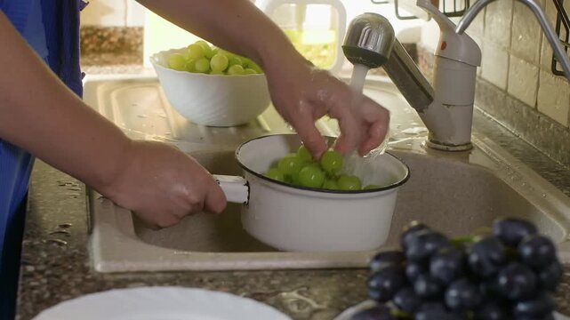 Person rinsing fresh green grapes in white colander at kitchen sink, bright natural light streaming through window, fruit preparation close up.