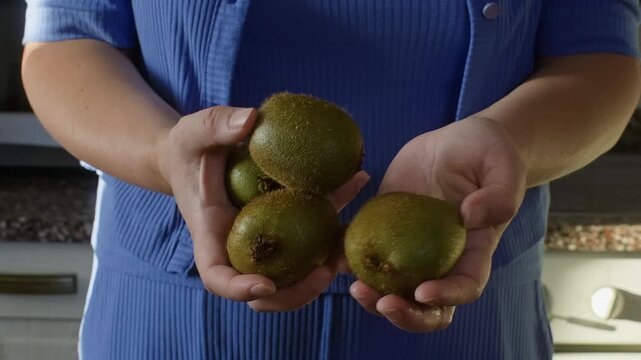 Adult woman in blue shirt displays fresh kiwis in cupped hands with slight camera movement, showcasing organic produce in bright indoor setting.