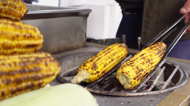 Close-up cinematic motion of a street vendor grilling corn on the cob over hot coals, turning it with tongs, fresh golden kernels with char marks on an outdoor market grill