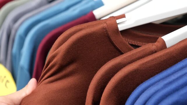 Close-up motion of hand selecting brown shirt on rack, showcasing variety of colorful garments in clothing store or boutique