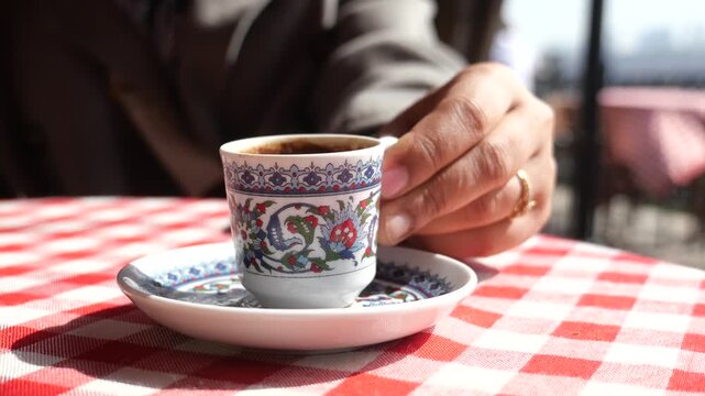 Person lifting small cup of traditional turkish coffee from saucer on checkered tablecloth at outdoor cafe