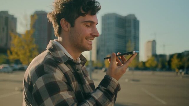 Happy Caucasian man recording voice message on smartphone while walking city street smiling guy businessman chatting mobile chat online sending audio voicemail using messenger speakerphone outdoors
