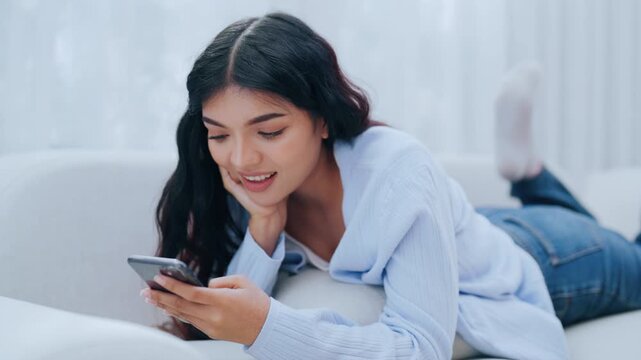 Young Asian woman using her cellphone while relaxing on the sofa at home, typing chat message on smartphone, smiling girl use cell phone chatting, searching information on browser wireless internet