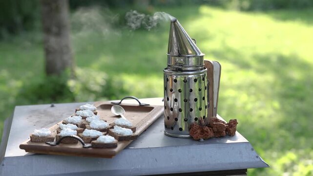 Metal beekeeping smoker releases smoke beside small bread pieces with white spread on a wooden tray, set outdoors against a sunlit green garden