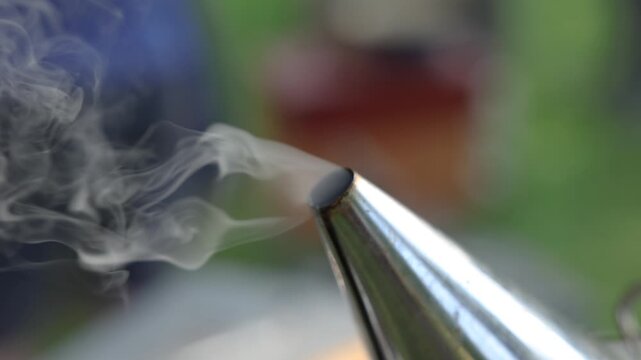 Close-up of a metal beekeeping smoker emitting curling white smoke outdoors, with a softly blurred green and brown background and shallow depth of field