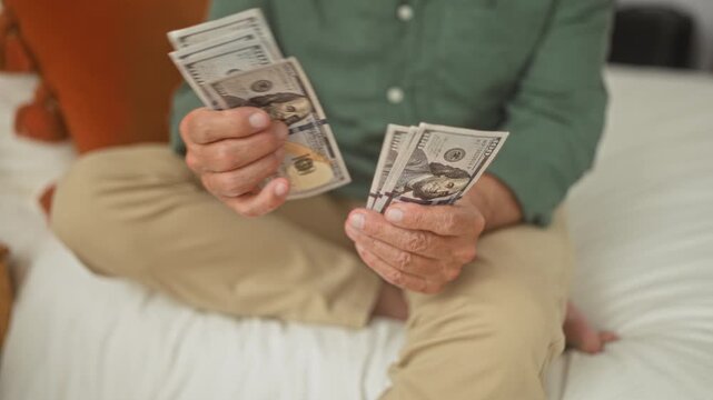 Man counting dollars on bed, hands fanning hundred dollar banknotes in a building bedroom; retirement savings relief.