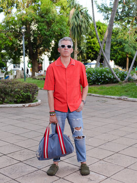 Man in Red Shirt and Ripped Jeans Posing with Handbag in a Public Park