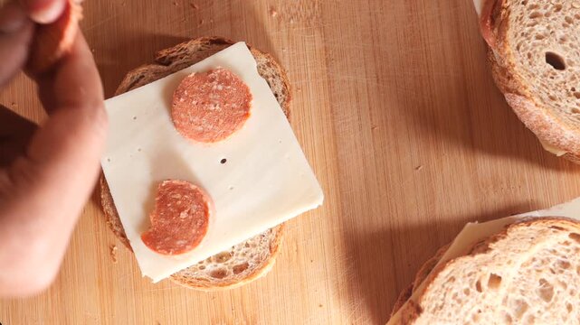 Making sandwiches from overhead shot, male hands put slices of pepperoni and cheese on a slice of bread on a wooden surface, ready to be cooked